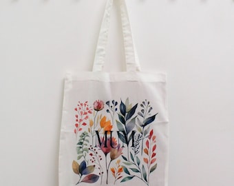 May include: White canvas tote bag featuring a floral design in shades of pink, orange, yellow, green, and blue. The word "MUM" is printed in black in the centre of the design.