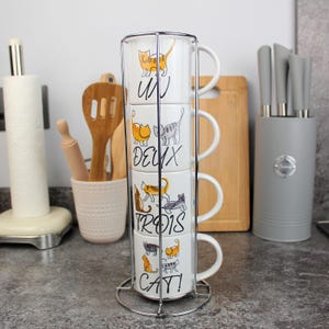 May include: A set of four white ceramic mugs stacked in a metal rack. Each mug features a different cat illustration and the numbers "UN", "DEUX", "TROIS", and "CAT!" in black script. The mugs have white handles.