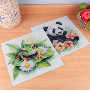 Puede incluir: Dos salvamanteles cuadrados de cerámica. Uno presenta una rana verde con ojos rojos y patas naranjas rodeada de margaritas blancas y hojas verdes. El otro representa un oso panda rodeado de flores rosas y naranjas.