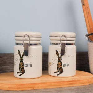 May include: Two cream ceramic storage jars with silver metal clasps and lids. Each jar has a brown hare illustration and the words "COFFEE" and "SUGAR". The jars are on a wooden board.