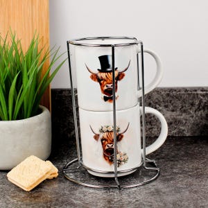 May include: Two white ceramic mugs stacked in a chrome stand. Each mug features a Highland cow illustration. The top mug has a cow wearing a top hat and bow tie. The bottom mug has a cow wearing a floral crown and necklace.