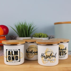 Peut inclure: Quatre bocaux en céramique blanche avec des couvercles en bambou, chacun représentant un type de noix différent : amande, noix, noisette et noix mélangées. Les bocaux ont du texte noir et des illustrations de noix.