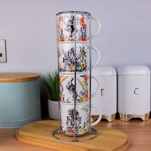Può includere: Un set di quattro tazze impilate in ceramica bianca con illustrazioni di Alice nel Paese delle Meraviglie, tenute in un supporto cromato. Le tazze presentano disegni floreali e di personaggi.
