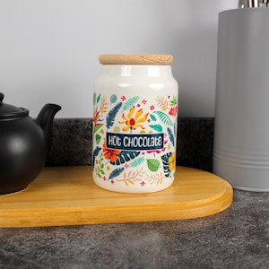 Op de afbeelding: Een witte keramische pot met een houten deksel, met het opschrift "Hot Chocolate". De pot is versierd met kleurrijke bloemen- en bladerpatronen in oranje, geel, blauw en groen. Hij staat op een houten dienblad, naast een zwarte theepot.