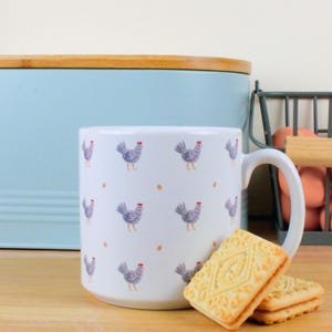 Può includere: Una tazza in ceramica bianca con un motivo di galline grigie e punti arancioni. La tazza ha un manico ed è appoggiata su una superficie di legno accanto a due biscotti.
