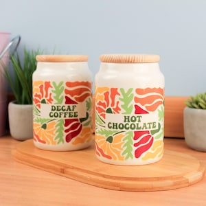 Op de afbeelding: Twee keramische potten met houten deksels, één met het opschrift "DECAF COFFEE" en de andere met "HOT CHOCOLATE". De potten zijn wit met een kleurrijk bloemmotief in oranje, rood en groen. Ze staan op een houten snijplank.