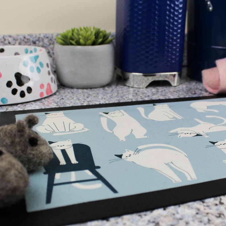 May include: A blue and white cat-themed kitchen mat with a black border. The mat features a variety of white cats in different poses on a light blue background.