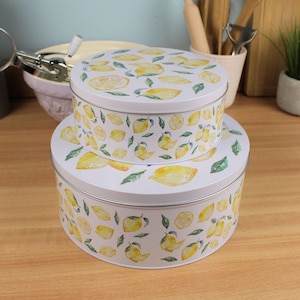 May include: Two white, round metal cake tins stacked. They feature a pattern of yellow lemons and green leaves. The background includes kitchen utensils and a small potted plant. The tins are ideal for storing baked goods.