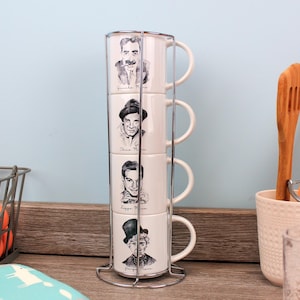 May include: A set of four white ceramic mugs featuring portraits of comedians, held in a silver stand. Kitchen utensils and a basket of oranges are also visible. The mugs are for tea or coffee.