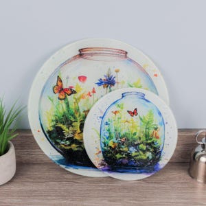 Puede incluir: Dos fuentes redondas decorativas con diseño de terrario. La fuente más grande tiene 30 cm de diámetro y la más pequeña 20 cm. Cada una presenta una colorida ilustración de mariposas y plantas.