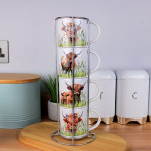 May include: Four white ceramic mugs with Highland cow illustrations, stacked in a metal rack. The mugs are on a wooden surface. The background includes a light blue bread bin and white canisters with gold bases. The mugs are for tea or coffee.