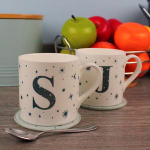 Può includere: Due tazze in ceramica bianca con un motivo a stelle blu e bianco. La tazza a sinistra ha la lettera "S" e la tazza a destra ha la lettera "J". Le tazze sono appoggiate su sottobicchieri abbinati blu e bianco.