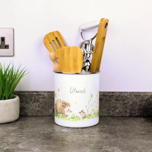 Könnte beinhalten: Ein weißer, zylindrischer Utensilienhalter aus Metall mit einem Aufdruck von einem Igel und einer Maus in einer Wiese mit gelben Blumen. Das Wort Utensils ist in schwarzer Schrift gedruckt. Er enthält Bambusspatel und einen Schneebesen.