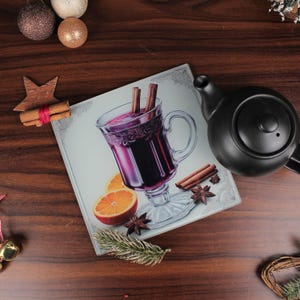 Può includere: Un tagliere decorativo in vetro con un motivo di vin brulé, decorato con bastoncini di cannella e fette d'arancia. Una teiera nera e decorazioni natalizie su una superficie di legno.