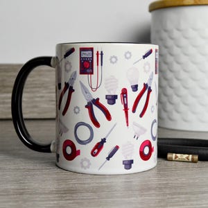 May include: White ceramic mug with a black handle. The mug has a design of various electrical tools, including pliers, screwdrivers, and a light bulb. The tools are in shades of red, blue, and gray.