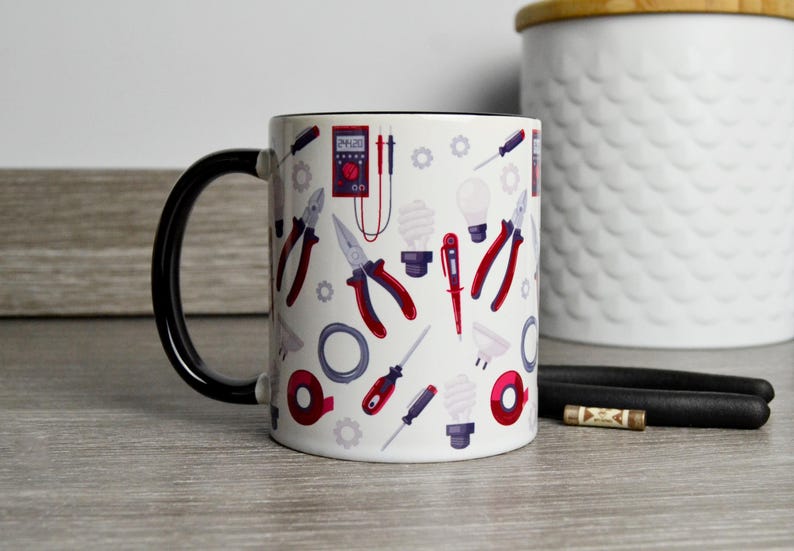 May include: White ceramic mug with a black handle. The mug has a design of various electrical tools, including pliers, screwdrivers, and a light bulb. The tools are in shades of red, blue, and gray.