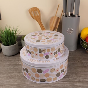 Peut inclure: Deux boîtes à biscuits rondes blanches empilées. Elles sont décorées d'illustrations de biscuits beiges, roses et bruns. Le texte "HANDS OFF MY BISCUITS!" est imprimé sur le dessus de chaque boîte.