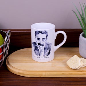 May include: A white ceramic mug featuring a dark blue watercolour-style portrait of Groucho Marx with his signature moustache, glasses, and cigar. The name Groucho Marx is printed in a cursive script directly below the illustrated portrait.