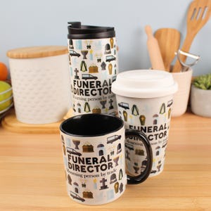 Puede incluir: Una taza de cerámica blanca, un vaso de viaje y un vaso con asa y tapa negros. Las tazas presentan un patrón repetido de iconos funerarios y el texto "FUNERAL DIRECTOR". La taza tiene un interior negro.