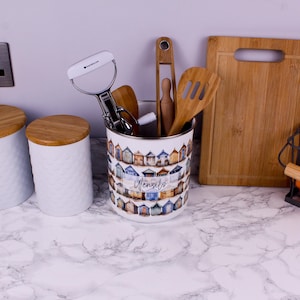 May include: A white cylindrical metal utensil holder featuring a repeating pattern of colourful beach huts and the word Utensils printed in script. The container holds a metal vegetable peeler, wooden tongs, and a wooden slotted spatula.