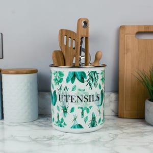 Op de afbeelding: Een witte cilindervormige metalen keukengereihouder met een groen botanisch patroon van diverse kruiden en het woord UTENSILS in zwarte letters. De houder is gevuld met diverse houten keukengerei, waaronder een spatel en een tang.