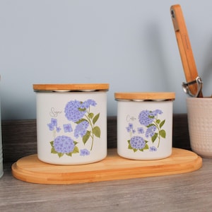 Può includere: Due barattoli in ceramica bianca con coperchi in legno, etichettati "Zucchero" e "Caffè". Ogni barattolo presenta un motivo floreale di ortensie viola. I barattoli sono su un vassoio di legno, suggerendo un contenitore da cucina.