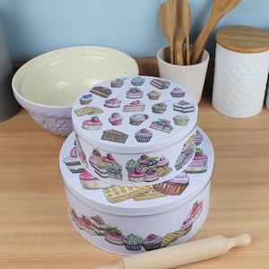 Peut inclure: Deux boîtes à gâteaux rondes en métal blanc empilées. Les couvercles sont décorés d'illustrations colorées de gâteaux, de cupcakes et de gaufres. Un rouleau à pâtisserie et d'autres articles de cuisine sont en arrière-plan.