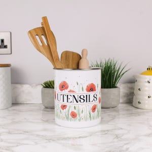 Puede incluir: Porta utensilios de cocina blanco con diseño de amapolas y la palabra "UTENSILS" en una pancarta roja. Contiene utensilios de cocina de madera. El fondo incluye una encimera de mármol y pequeñas plantas en macetas.
