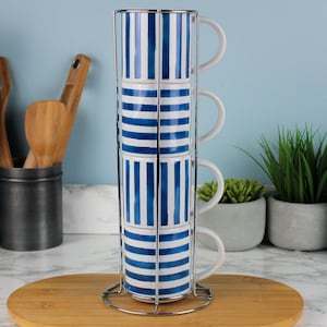 May include: A set of four stacked white ceramic mugs with blue vertical stripes, held in a chrome wire rack. The mugs have white handles and are displayed on a wooden serving board. The background is a light blue wall.