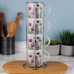 Peut inclure: Un ensemble de quatre tasses en céramique blanche empilées dans un support en métal. Chaque tasse est décorée d'un motif floral avec des fleurs roses, violettes et bleues. Les tasses sont sur un plateau en bois, avec des ustensiles de cuisine en arrière-plan.