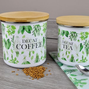 May include: Two white canisters with wooden lids, labelled "Decaf Coffee" and "Decaf Tea." The canisters are decorated with green botanical illustrations. A small pile of coffee granules sits near the coffee canister.