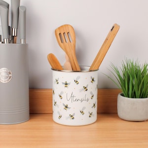 Könnte beinhalten: Weißer Emaille-Küchenutensilienhalter mit Bienenmuster und dem Wort "Utensils". Er enthält Holzlöffel und -spatel. Ein grauer Messerblock und eine kleine Topfpflanze sind ebenfalls zu sehen.