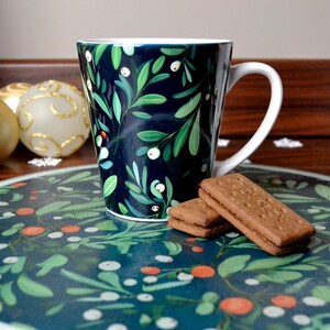 May include: A white ceramic mug with a dark blue and green floral pattern. The mug is sitting on a round plate with a matching pattern. Two brown biscuits are on the plate.