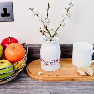 May include: A white ceramic vase with a pink floral design holds flowering branches. A white mug and a wooden tray with biscuits are next to the vase. A metal fruit basket with apples, oranges, and pears is on the left.