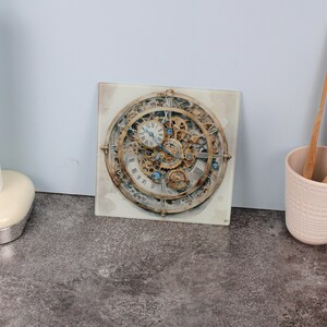 Peut inclure: Tableau carré représentant un design d'horloge détaillé avec des engrenages apparents et des chiffres romains. Le cadran de l'horloge a une esthétique vintage avec des accents dorés, bronze et bleus. L'œuvre est montée sur un fond carré blanc cassé.