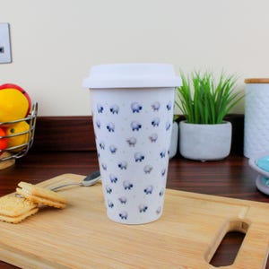 May include: White ceramic travel mug with a white lid. The mug features a repeating pattern of blue and white sheep.