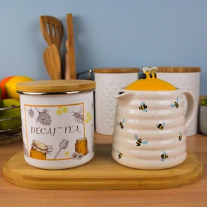 Peut inclure: Une théière en céramique blanche avec un couvercle jaune et des motifs d'abeilles, à côté d'un pot blanc avec un couvercle en bois. Le pot porte une étiquette "Decaf Tea" avec des illustrations d'abeilles et de miel. Les deux sont sur un plateau en bois.