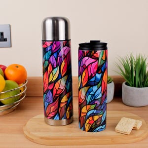 Op de afbeelding: Een roestvrijstalen thermoskan- en reismokset. Beide items hebben een kleurrijk glas-in-lood bladontwerp in tinten rood, oranje, geel, paars en blauw. De set wordt gepresenteerd op een houten snijplank.