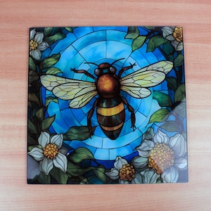 Puede incluir: Azulejo decorativo cuadrado con un diseño de abeja estilo vidriera. La abeja es marrón y amarilla, sobre un fondo azul con detalles florales y de hojas. Flores blancas con centros amarillos y hojas verdes enmarcan la abeja.