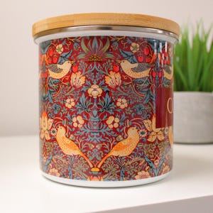 May include: A ceramic canister with a wooden lid features a colorful floral and bird pattern. The pattern includes red, blue, green, yellow, and white colors. The canister is labeled "Coffee".