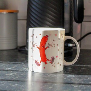 May include: White ceramic mug featuring a red and white cartoon sausage character with stars in the background. The sausage is smiling and has arms and legs.