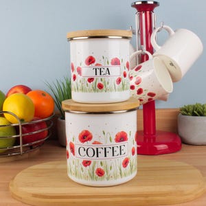 Könnte beinhalten: Weiße Emaille-Vorratsdosen mit Holzdeckeln, beschriftet mit „TEA“ und „COFFEE“, verziert mit roten Mohnblumen. Ein roter Tassenbaum hält weiße Tassen. Ein Drahtkorb mit Obst und eine kleine Topfpflanze sind ebenfalls zu sehen.