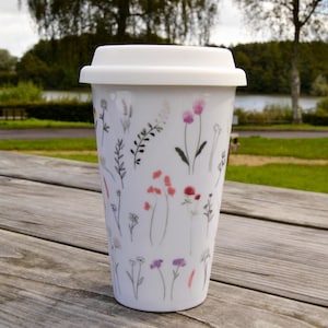Könnte beinhalten: Ein weißer Keramik-Reisebecher mit Silikondeckel, der mit einem zarten botanischen Muster aus verschiedenen Wildblumen in Rosa-, Lila- und Grautönen verziert ist. Der Becher hat eine konische zylindrische Form und eine glatte Oberfläche mit floralen Motiven.