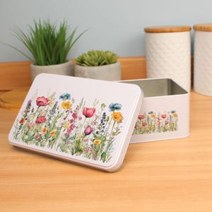 Op de afbeelding: Een rechthoekige witte metalen opbergdoos met een bloemenontwerp met rode, roze, gele en blauwe bloemen. Het deksel staat een beetje open en onthult de binnenkant. De doos staat op een houten oppervlak.