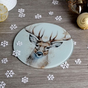 May include: A round glass trivet with a blue background and a watercolour illustration of a deer with antlers. The trivet is surrounded by white snowflakes.