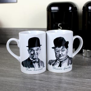 Könnte beinhalten: Zwei weiße Keramikbecher mit Schwarzweißbildern von Stan Laurel und Oliver Hardy. Die Becher haben schwarze Henkel und der Text "Stan Laurel" und "Oliver Hardy" ist auf der Unterseite jedes Bechers aufgedruckt.