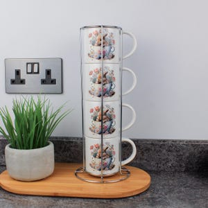 Könnte beinhalten: Ein Set aus vier gestapelten weißen Keramikbechern mit Blumen- und Hasenmuster, gehalten in einem Chrom-Metallgestell. Die Becher sind in Pastellfarben gehalten. Das Set steht auf einem Holztablett. Die Becher sind ca. 10 cm hoch.