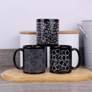 May include: Three black ceramic mugs with white geometric patterns are displayed on a wooden tray. One mug features a vertical dash design, another a square pattern, and the third a curved line design. The mugs are for beverages.