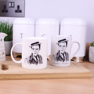 Puede incluir: Dos tazas de cerámica blanca con un retrato en blanco y negro de Buster Keaton. Las tazas tienen un asa y una base dorada. Una taza tiene el nombre "Buster Keaton" escrito en cursiva.