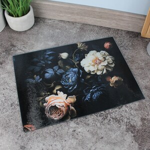 May include: Rectangular glass chopping board with a dark background and a floral design. The flowers are in shades of white, blue, and peach. A kitchen accessory.